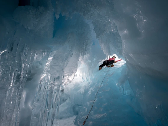 A climber is seen from below through a small opening in a mass of ice.