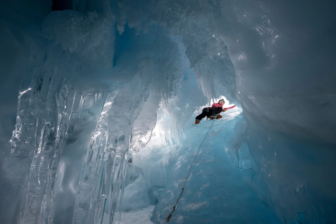 A climber is seen from below through a small opening in a mass of ice.